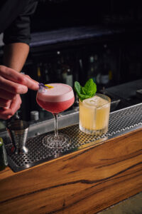 2 x cocktails on the bar. Pink cocktail left, yellow on left.